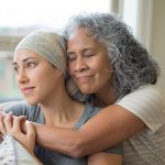 Hawaiian woman in 50s embracing her mid-20s daughter on couch who is fighting cancer